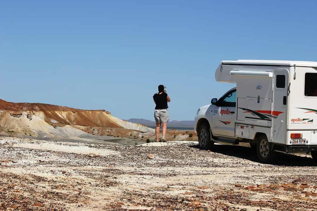 A man standing next to a 4WD campervan, looking out at a beautiful view, surrounded by blue sky.