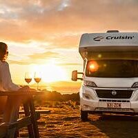 01 Cruisin Deluxe 4 Berth Min Sunset with a woman enjoying wine on outdoor terrace next to a campervan in Australia.