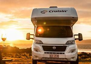 01 Cruisin Deluxe 4 Berth Min Sunset with a woman enjoying wine on outdoor terrace next to a campervan in Australia.
